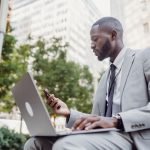 Businessman in a suit using a laptop and smartphone in an urban setting, blending technology with business. — Best Trading In